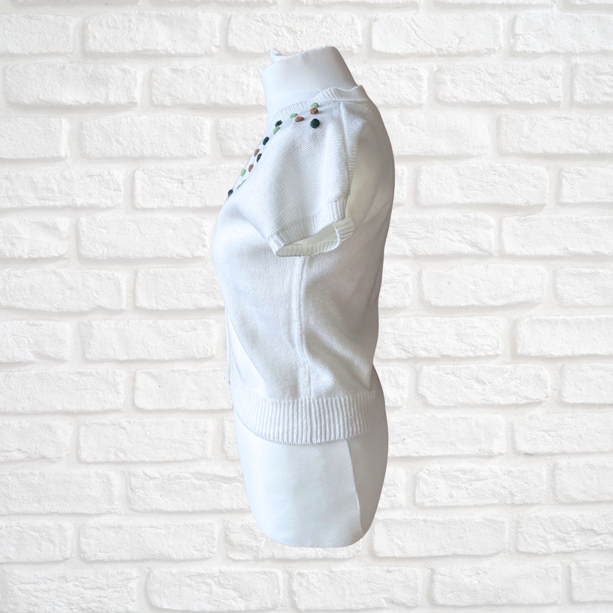A white knit sweater displayed on a white mannequin torso against a white painted brick wall. The sweater features small colorful embroidered details along the shoulder and a ribbed hem.