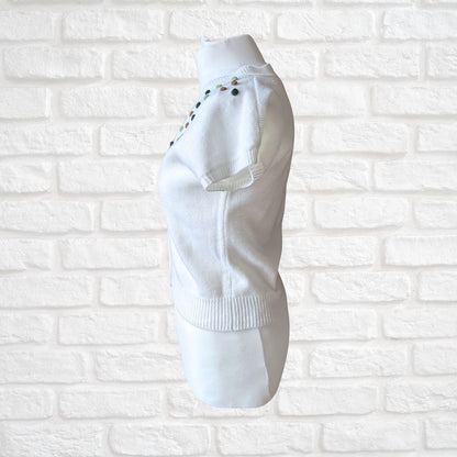A white knit sweater displayed on a white mannequin torso against a white painted brick wall. The sweater features small colorful embroidered details along the shoulder and a ribbed hem.