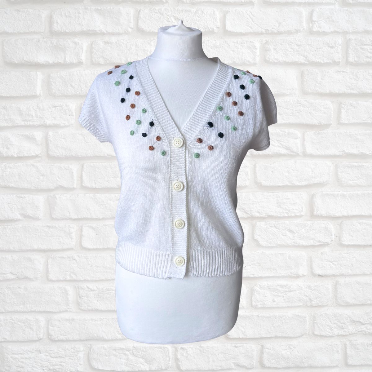 A white short-sleeve knit cardigan sweater displayed on a white mannequin torso against a white brick wall. The cardigan features a V-neck, front buttons, and colorful knit polka dots in green, brown, and black along the shoulders and upper chest.