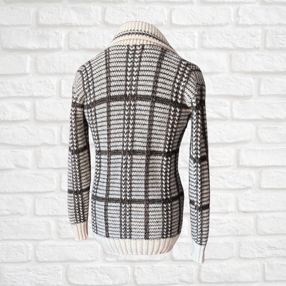 A long-sleeve knit sweater shown from the back, featuring a black and off-white plaid pattern with a ribbed cream collar, cuffs, and hem, displayed against a white brick wall background.