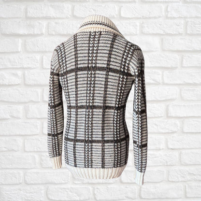 A long-sleeve knit sweater shown from the back, featuring a black and off-white plaid pattern with a ribbed cream collar, cuffs, and hem, displayed against a white brick wall background.
