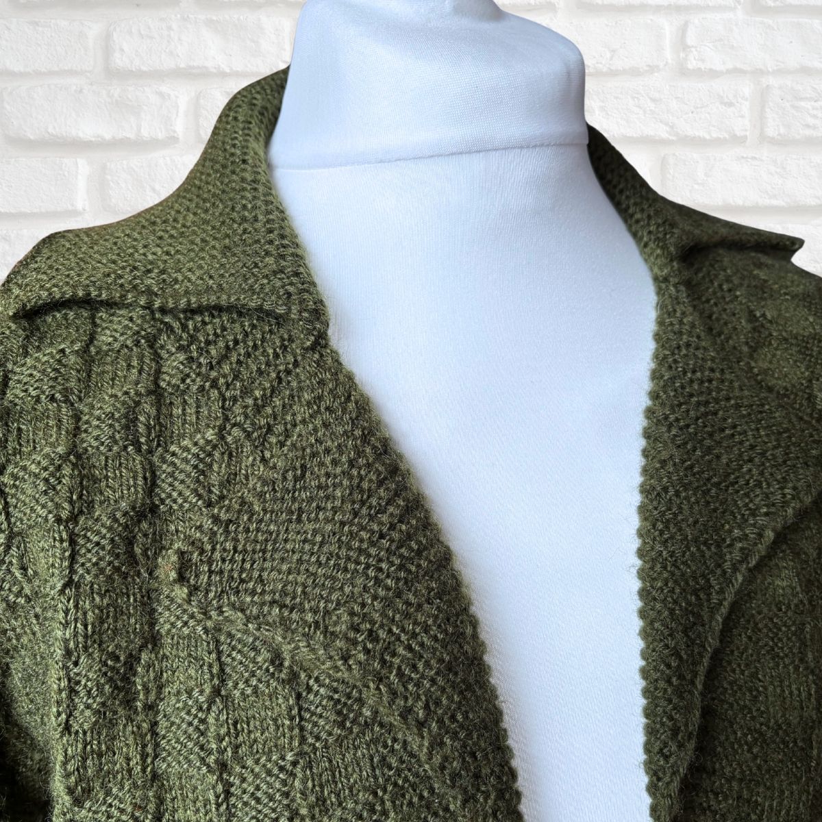 An olive green cable-knit cardigan sweater displayed on a white mannequin torso against a white brick wall background.