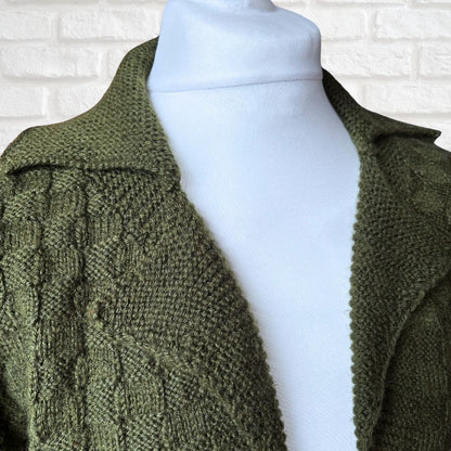 An olive green cable-knit cardigan sweater displayed on a white mannequin torso against a white brick wall background.