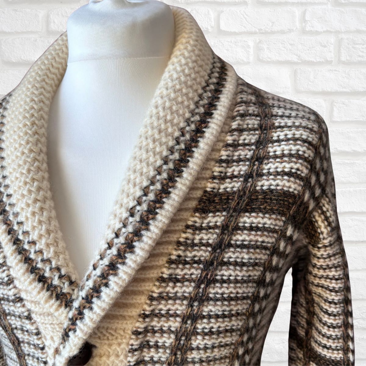 A close-up of a knit cardigan sweater displayed on a white mannequin torso against a white brick wall. The cardigan features a thick cream-colored shawl collar and a textured body with horizontal stripes in cream, brown, and black.
