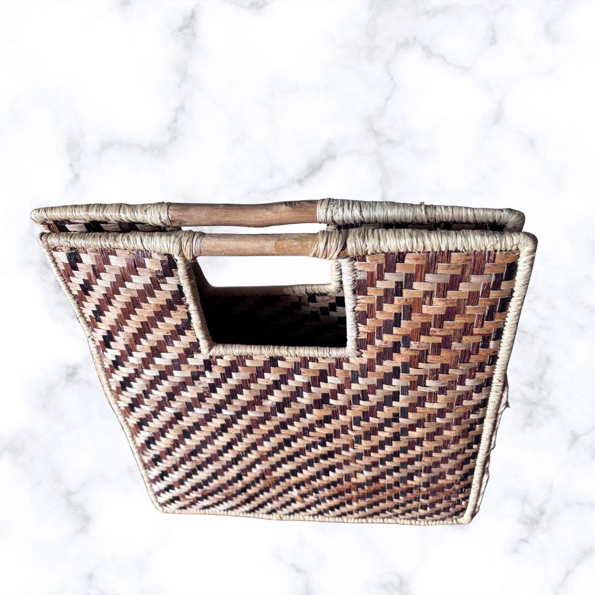 A woven straw basket with a rectangular cutout and a natural wood handle, shown from a top-down view on a white marble background.