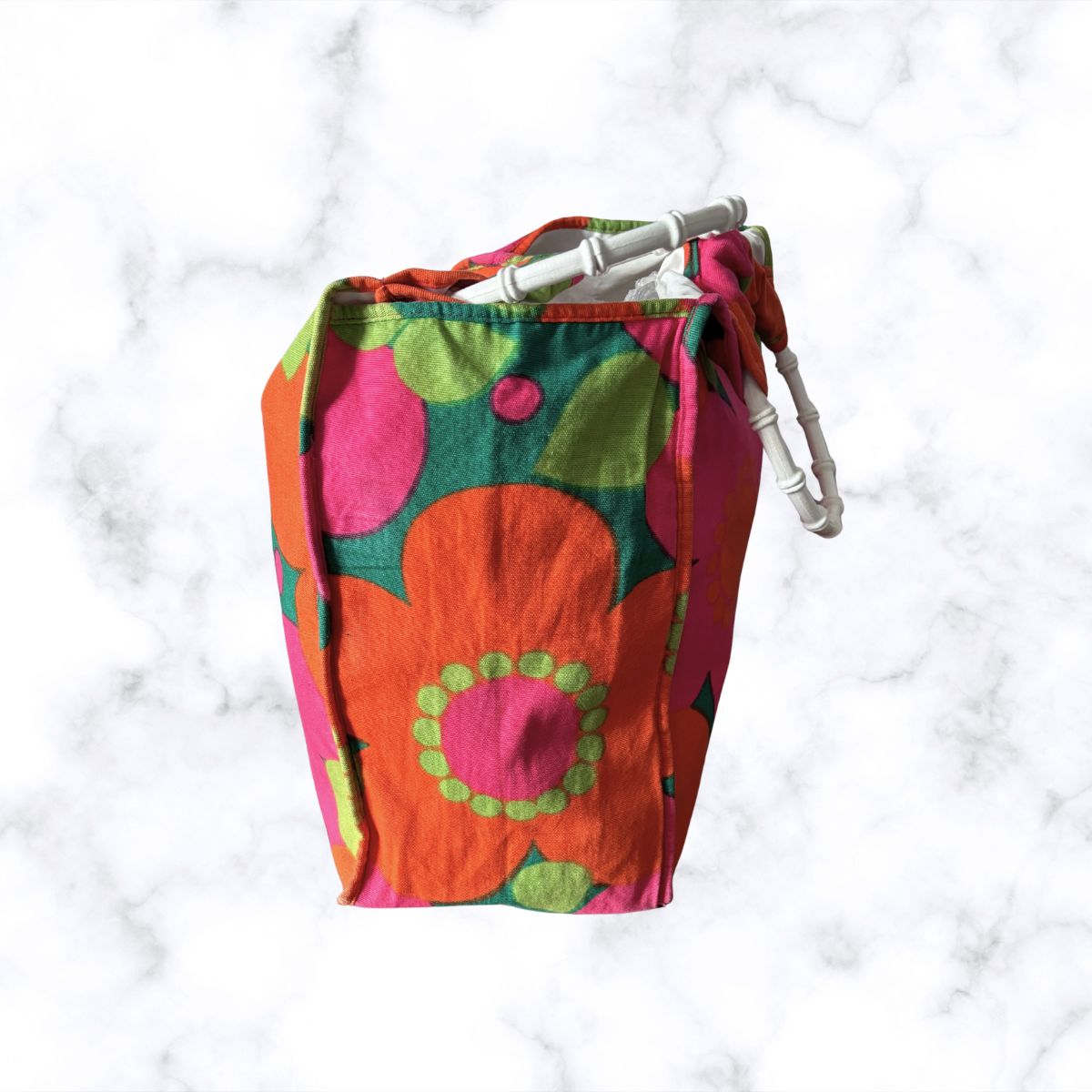 A fabric bag with a retro floral pattern in orange, pink, and green, featuring white plastic carabiner clips on the top, set against a white marble background.