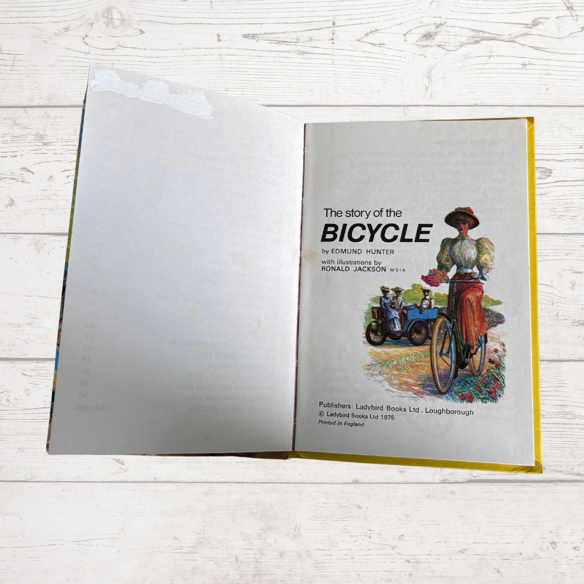 An open vintage book titled 'The story of the BICYCLE' lying on a white painted wood surface. The right page shows a colorful illustration of a person riding a bicycle, with publisher text at the bottom and a yellow border around the page.