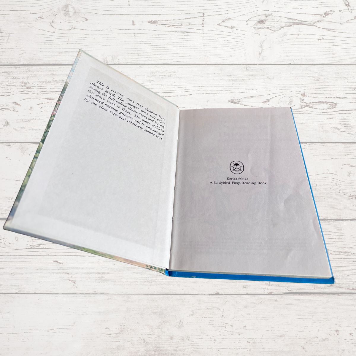 An open hardcover book lying on a white wood plank surface. The left page has printed text, and the right page shows a small circular logo
