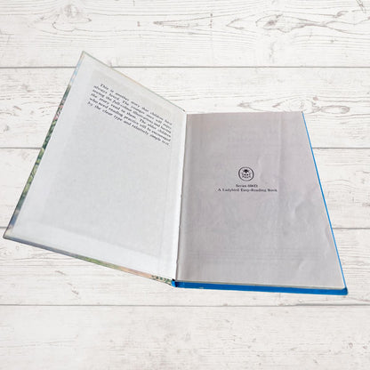 An open hardcover book lying on a white wood plank surface. The left page has printed text, and the right page shows a small circular logo